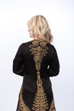 The image shows the Gwalior jacket. This is a highly embellished satin jacket that can be worn as a duster. The us extensive gold embroidery with red highlights on the back and sleeves of the of the jacket 