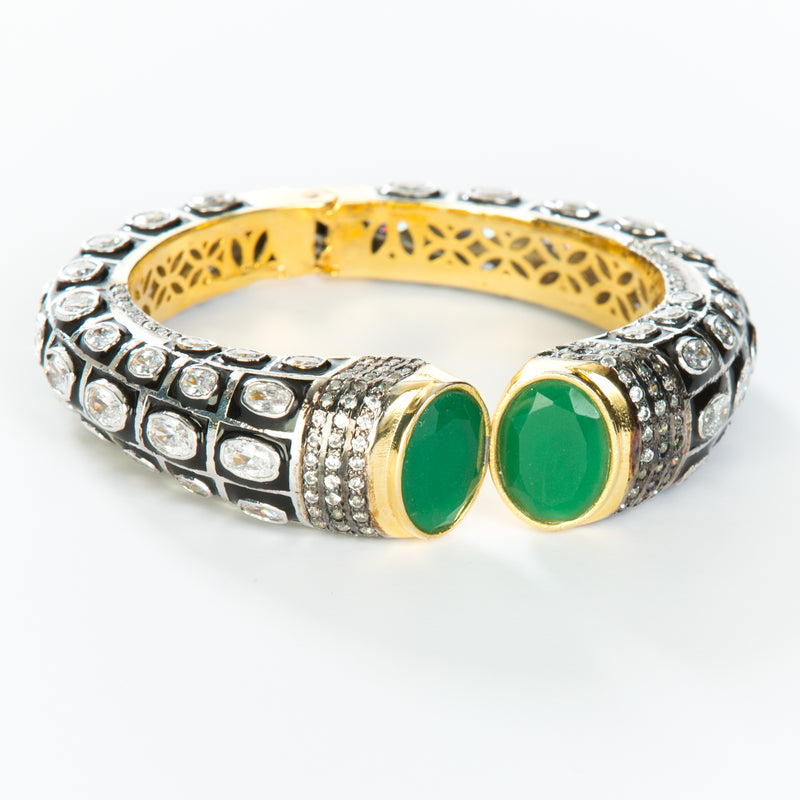 The images shows the Yatra.shop signature cuff. It is an easy to wear cuff with a hinge in the center. This cuff fits most wrists. It is embellished with enamel, Swarovski crystals of varying shapes and sizes and there are two round green semi precious stones at the opening. 