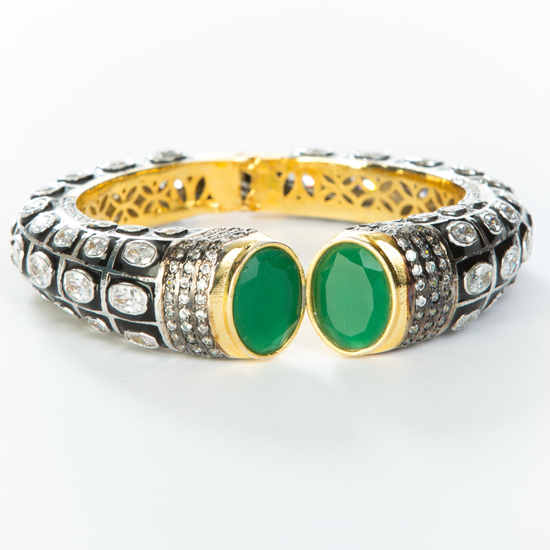 The images shows the Yatra.shop signature cuff. It is an easy to wear cuff with a hinge in the center. This cuff fits most wrists. It is embellished with enamel, Swarovski crystals of varying shapes and sizes and there are two round green semi precious stones at the opening. 