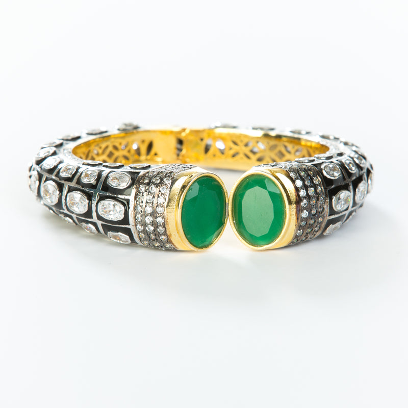 The images shows the Yatra.shop signature cuff. It is an easy to wear cuff with a hinge in the center. This cuff fits most wrists. It is embellished with enamel, Swarovski crystals of varying shapes and sizes and there are two round green semi precious stones at the opening. 