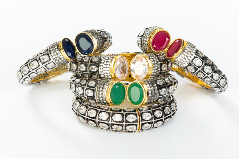 The images shows the Yatra.shop signature cuff. It is an easy to wear cuff with a hinge in the center. This cuff fits most wrists. It is embellished with enamel, Swarovski crystals of varying shapes and sizes and there are two round green semi precious stones at the opening. 